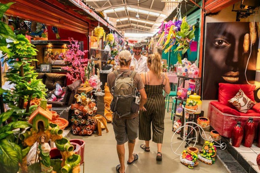 Un étroit couloir de marché bordé de différents étals où sont vendus des articles colorés, comme des plantes, de la poterie et des textiles. 1