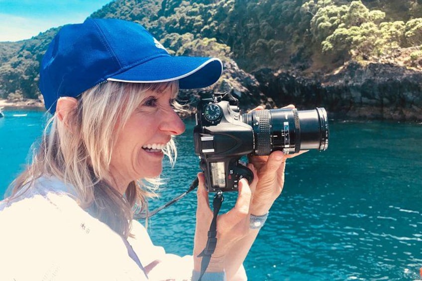 A person holding a camera with a lens, standing by a body of water with a rocky coastline in the background. 3