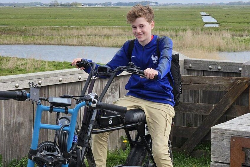 A person sitting on a blue electric bike with a camera mounted on the handlebars, in a rural setting with a wooden fence and a body of water in the background. 1