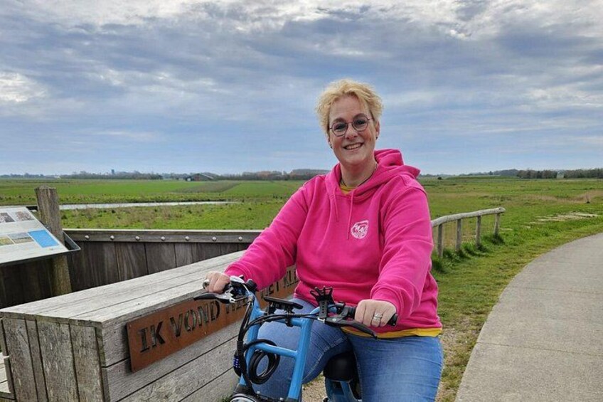 A person on a blue bicycle near a wooden sign with text, in a grassy area with a cloudy sky. 5