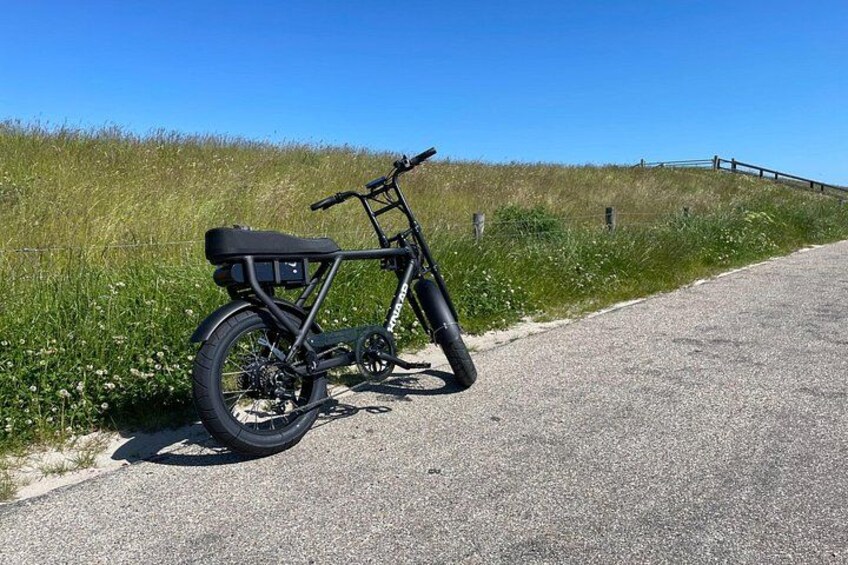A stationary electric bicycle parked on a paved path beside a grassy area with a fence in the background. 3