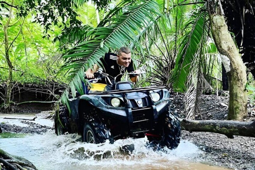 Una persona guida un quad attraverso un tratto d'acqua poco profondo, circondato da fitta vegetazione tropicale. 1
