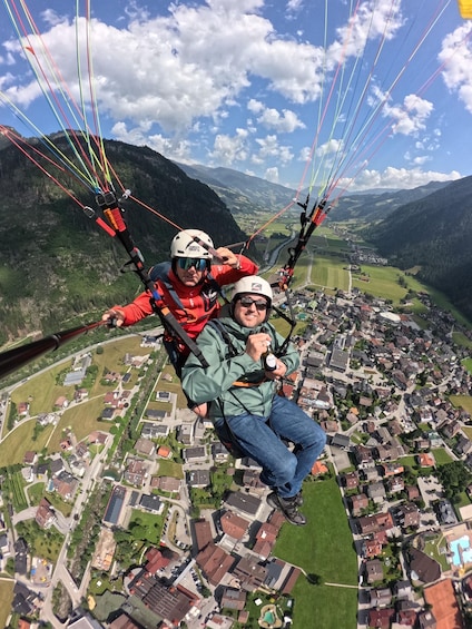 Zwei Personen gleiten mit einem Paragleiter über einer Stadt, im Hintergrund Berge. 5