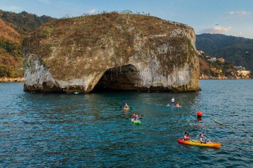 A large rock formation with a cave-like opening, kayakers on the water, and a mountainous landscape in the background. 1