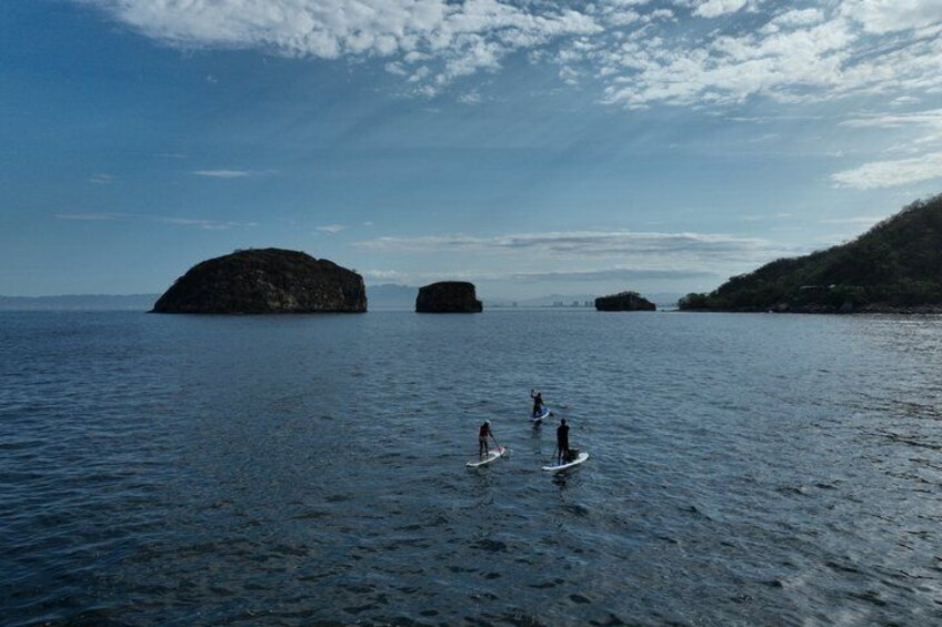 A calm sea with three paddleboarders, distant rocky outcrops, and a clear sky. 5
