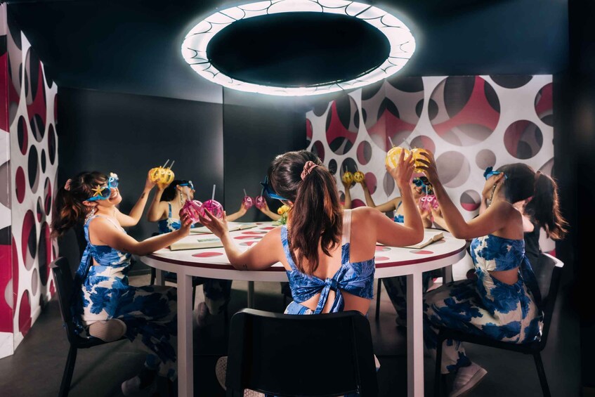 A group of people sitting around a round table, wearing blue floral dresses, in a room with a circular light fixture and patterned walls. 4