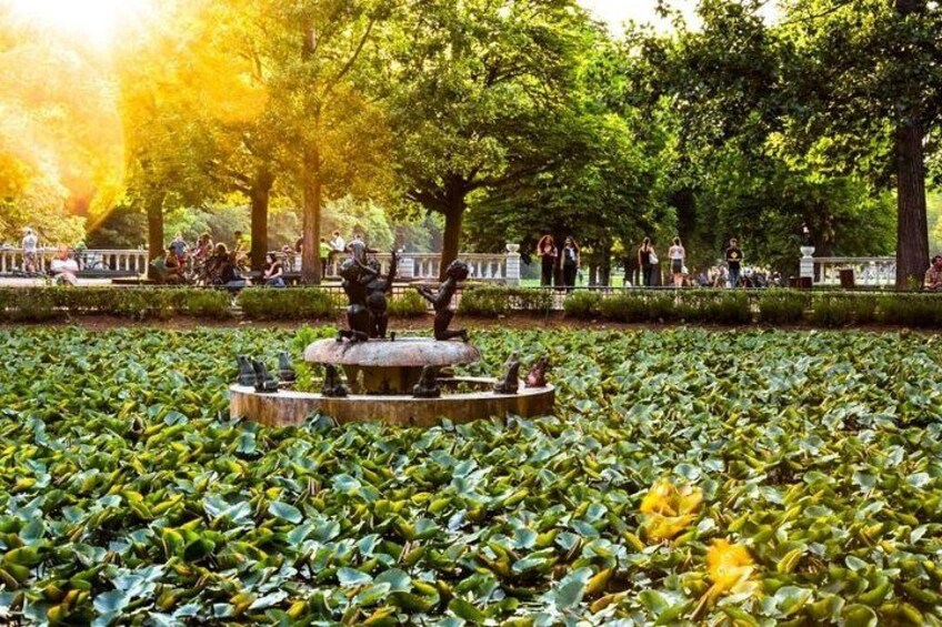 En park med en fontän omgiven av näckrosor, träd och människor. 1