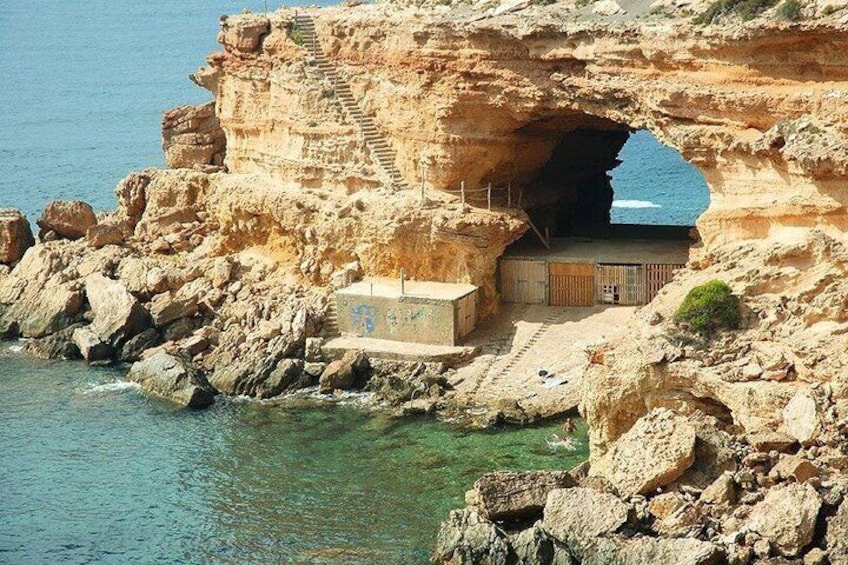 L’entrée d’une grotte côtière, avec un escalier menant à l’intérieur, est entourée d’un paysage rocheux et bordée par la mer. 2