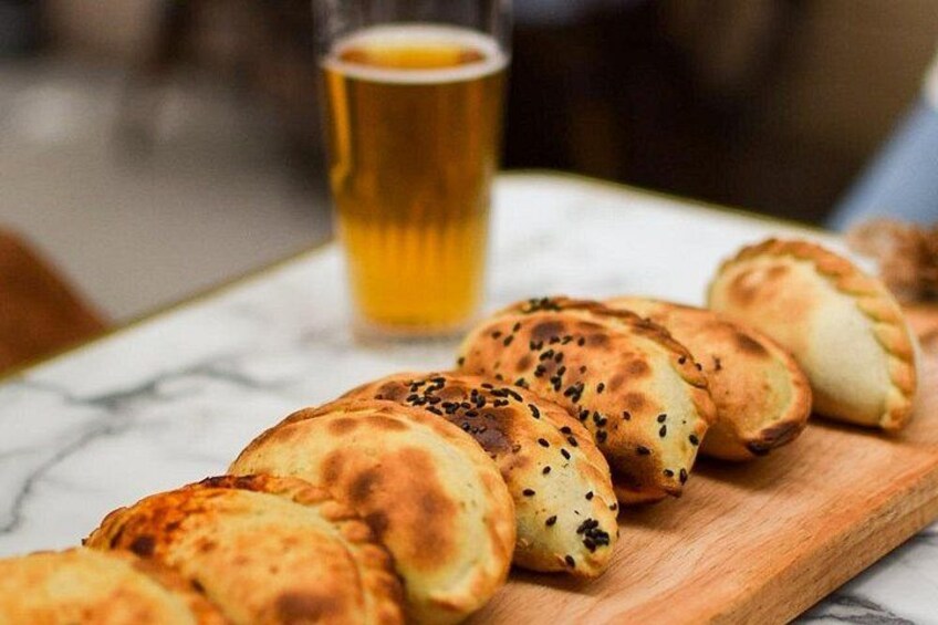 A wooden board with several round, golden-brown pastries topped with black sesame seeds, accompanied by a glass of amber liquid, likely beer, on a marble surface. 4