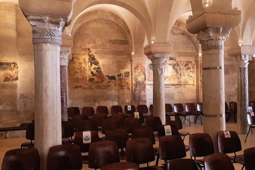 Ein Innenraum mit Steinsäulen, Bögen und einem Wandgemälde. Sitzreihen mit Stühlen sind angeordnet. 3