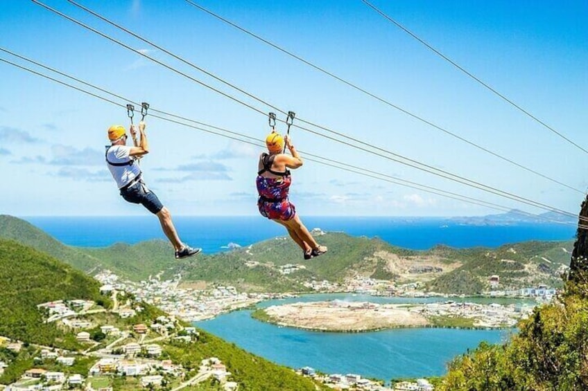 Two people ziplining over a coastal landscape with houses and a body of water below. 1