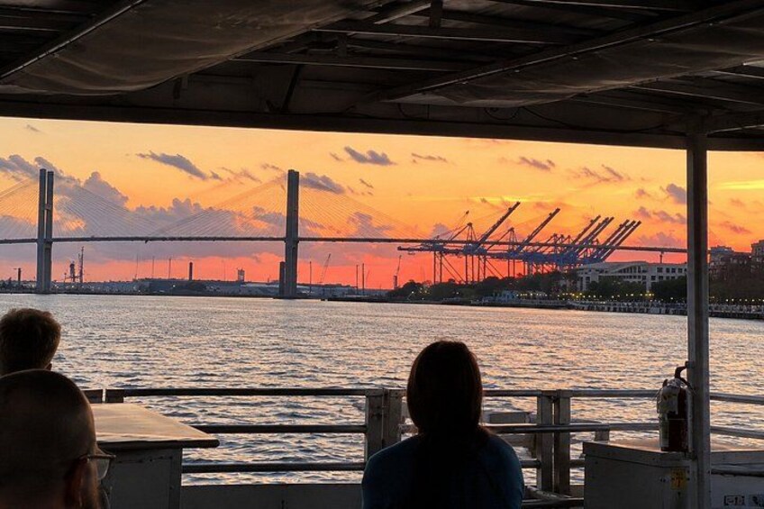 Vista del atardecer desde un bote, con un puente y grúas al fondo. 2