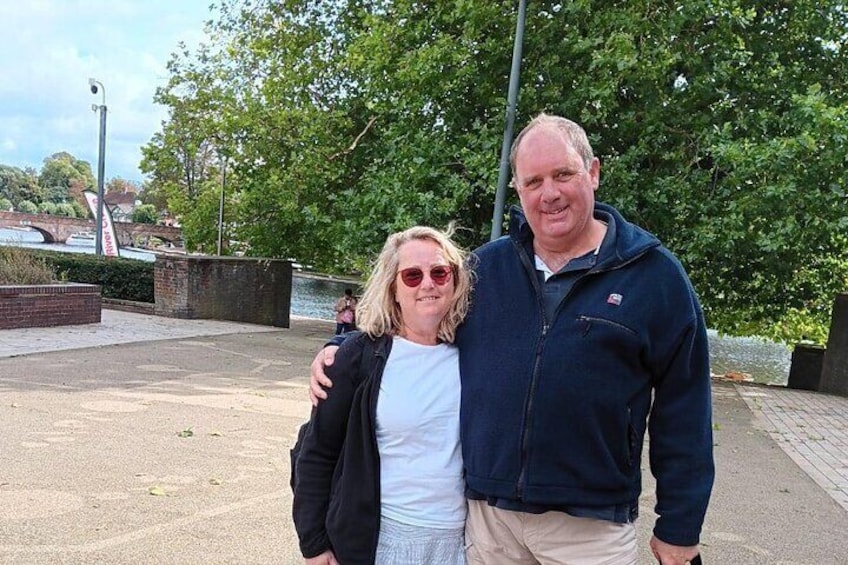 Two people standing on a paved area with a river and greenery in the background. 1