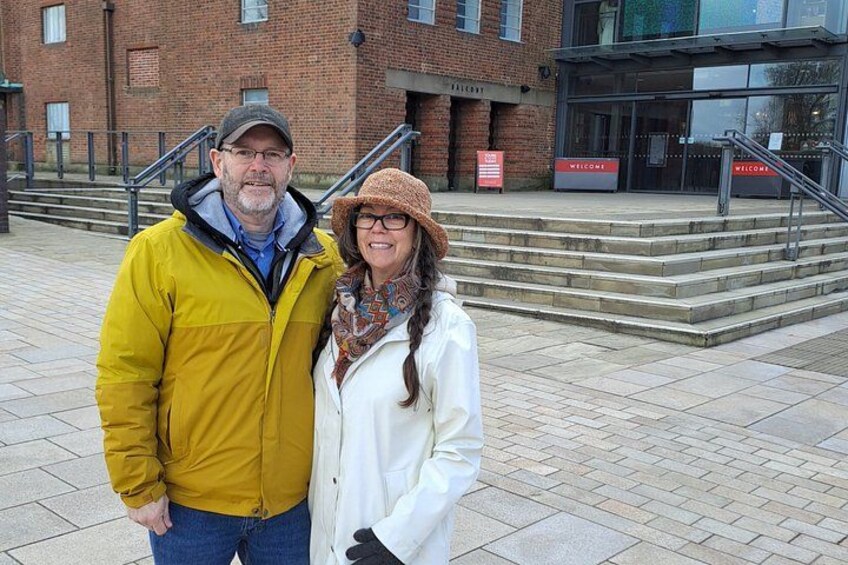 A man and a woman standing on a paved area with a brick building and a staircase in the background. 2