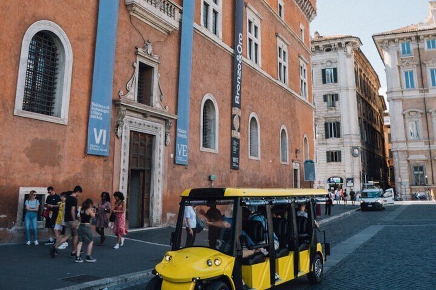 Un taxi amarillo en una calle adoquinada, personas de pie junto a un edificio con letreros 'Bronx' y 'VE', y un letrero 'VI' en el edificio. 3