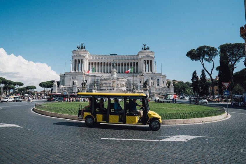 A yellow trolley on a cobblestone road, a large white building with columns and flags, and a roundabout with trees and people. 1