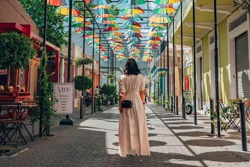 A woman in a long dress walking down a street with colorful umbrellas overhead. 1