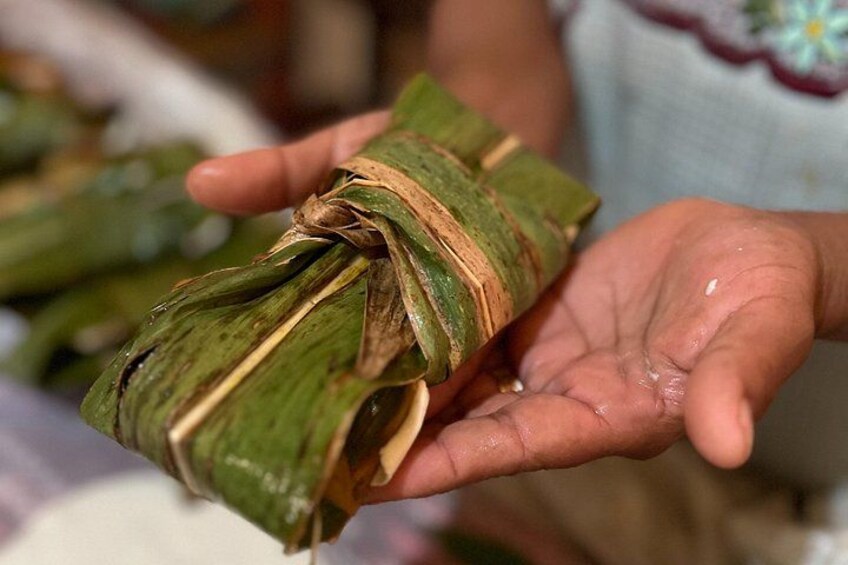 A person holding a wrapped food item, likely a traditional dish, with a green leaf wrapping and a brown string. 2