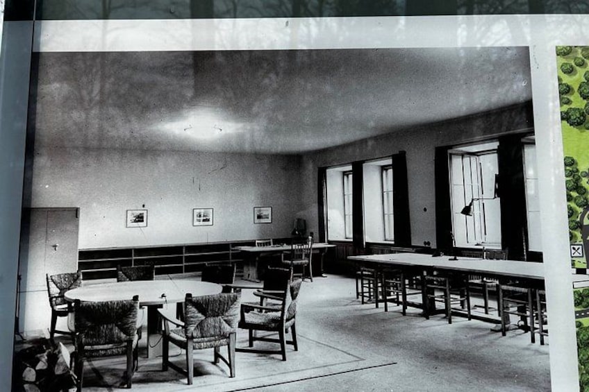 A classroom with long tables and chairs, framed pictures on the wall, and a window. 4