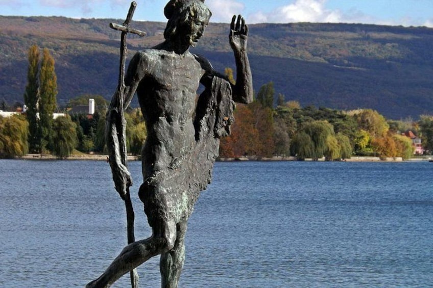 Eine Statue einer Figur, die eine Stange und ein Kreuz hält, steht an einem Gewässer vor einer bergigen Landschaft. 4