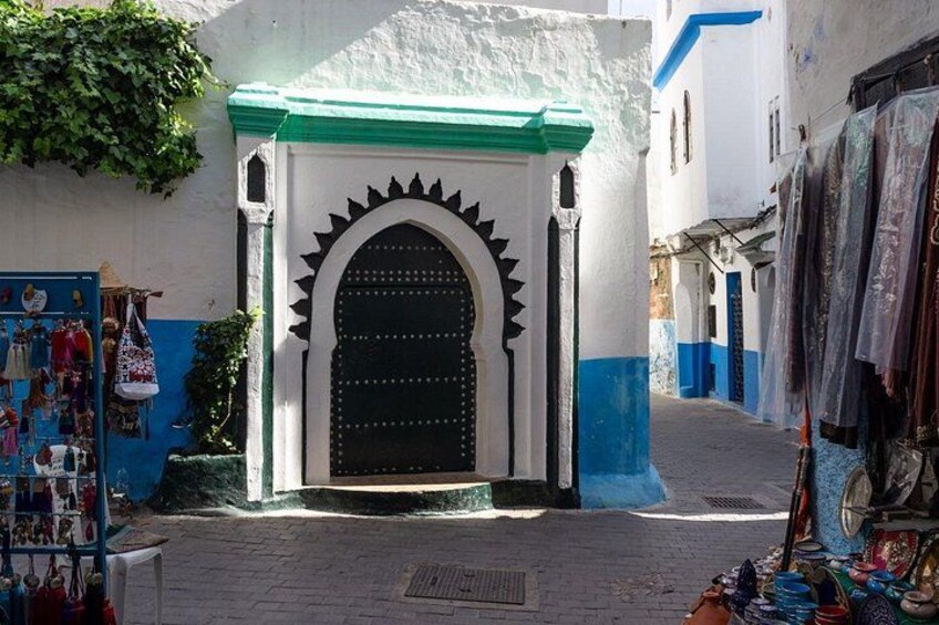 Una calle estrecha con un edificio azul y blanco, una puerta negra con un arco decorativo y una tienda con artículos de colores a la vista. 2