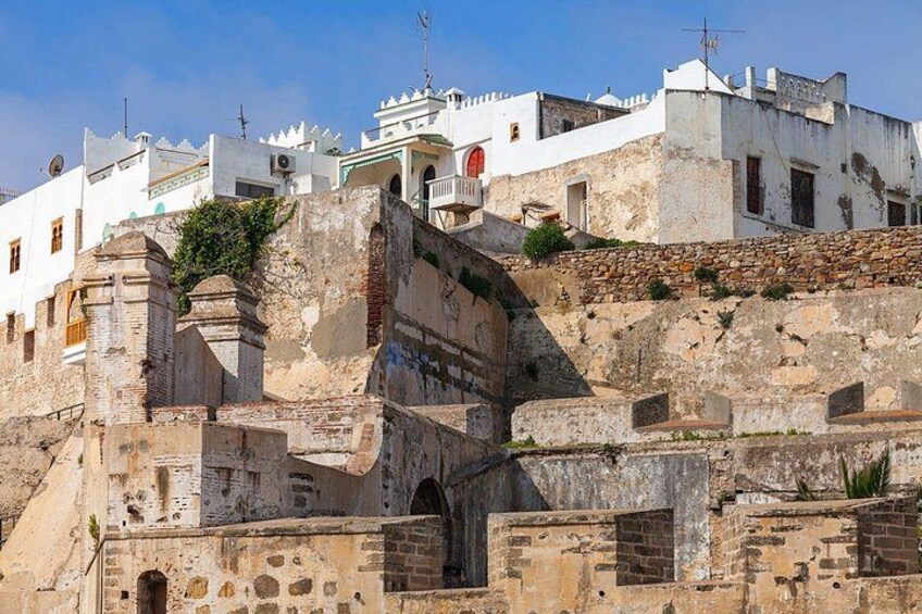 A historic fortified structure with stone walls and a gate, surrounded by white buildings with red accents. 3