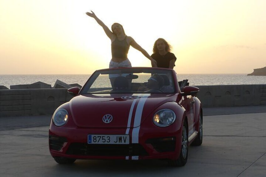 Une Volkswagen rouge décapotable, avec deux personnes debout sur le toit, et un coucher de soleil sur la côte en arrière-plan. 1
