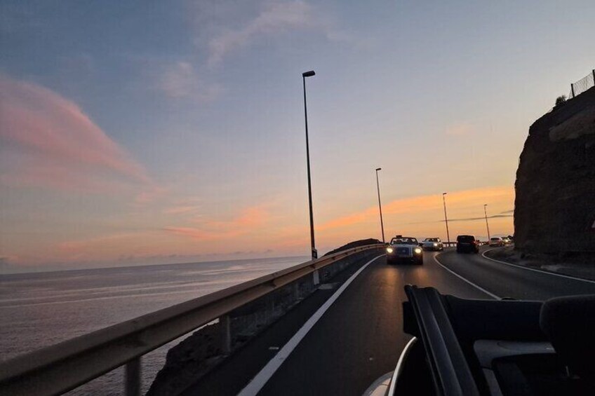 A coastal highway during sunset with vehicles, guardrails, and a rocky cliff. 2