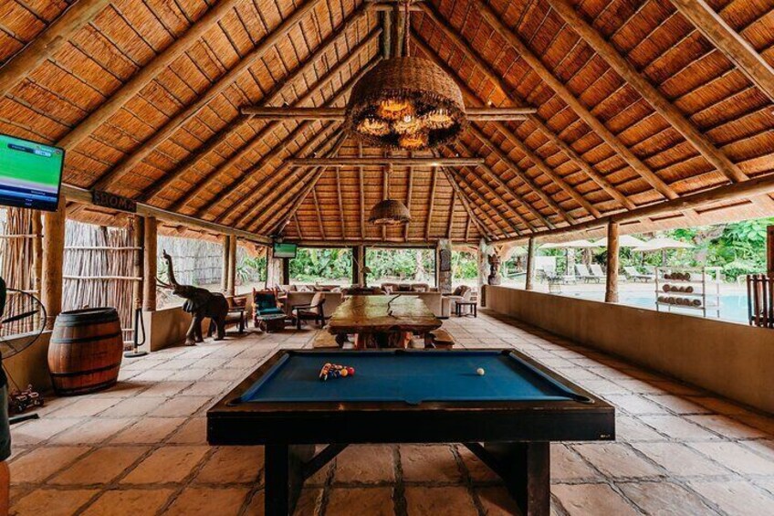 A pool table in a covered outdoor area with a thatched roof, wooden furniture, and a hanging light fixture. 4