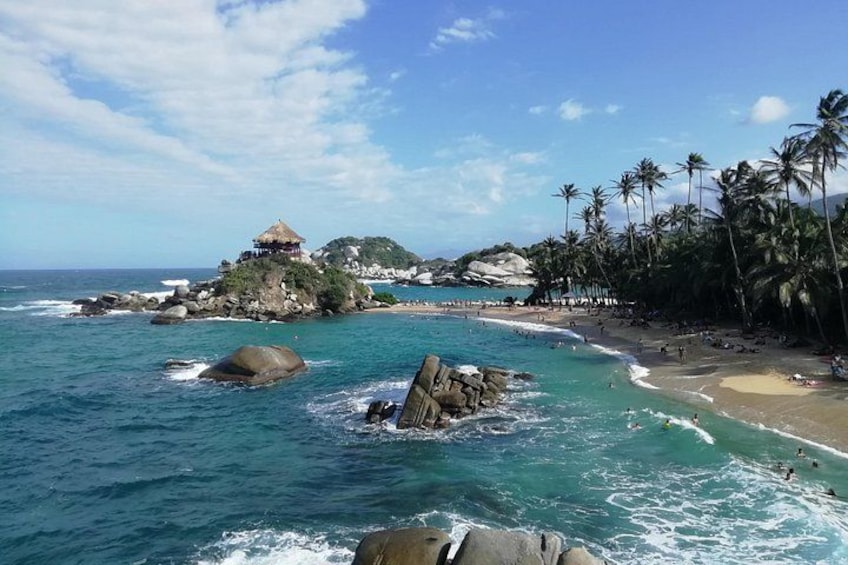 A tropical beach with clear turquoise waters, rocky outcrops, palm trees, and a small hut on a cliff. 1