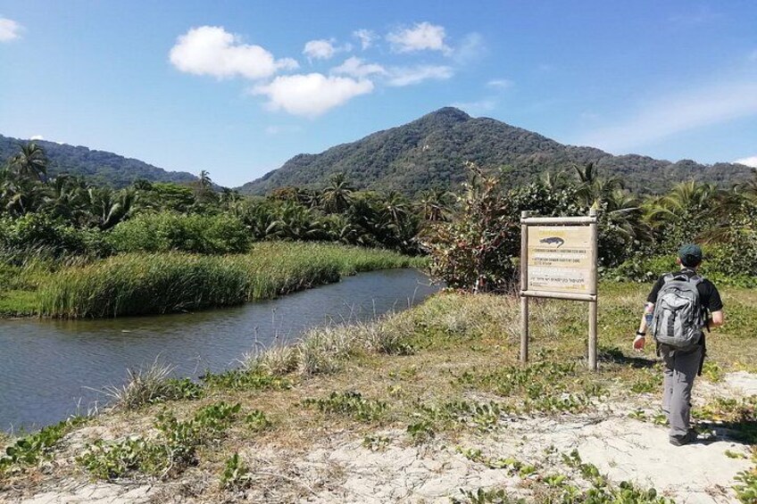 A river, a person with a backpack, and a signboard in a natural landscape with mountains in the background. 3