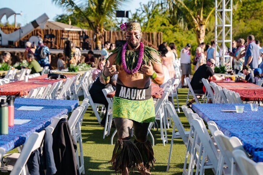 A person in a Hawaiian shirt and lei, walking through a crowded outdoor event with tables and chairs. 2