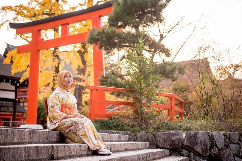A person in traditional attire sitting on stone steps, with a red torii gate and autumn foliage in the background. 1