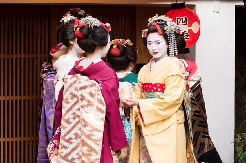 Geisha in traditional Japanese attire, including kimonos and headpieces, standing in front of a wooden building. 2