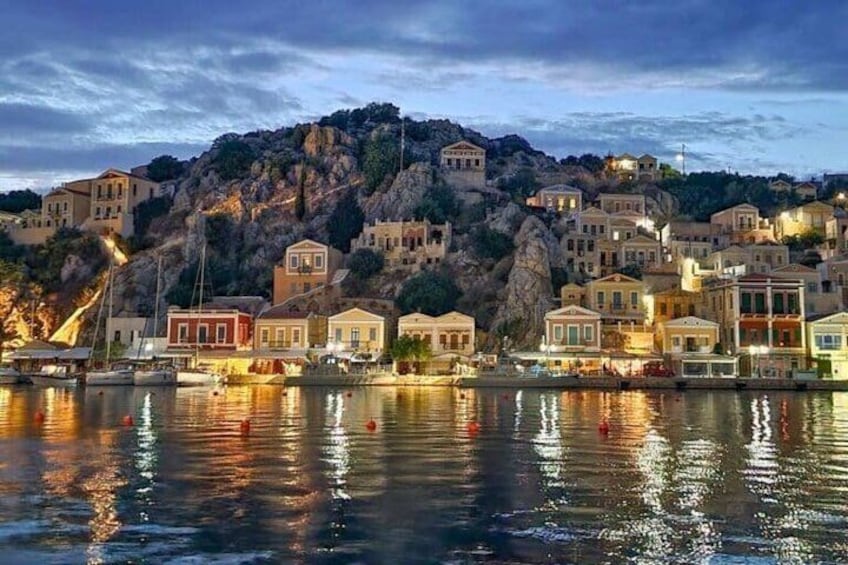 A coastal town at dusk with buildings reflecting on the water, a rocky hill in the background, and a clear sky. 2