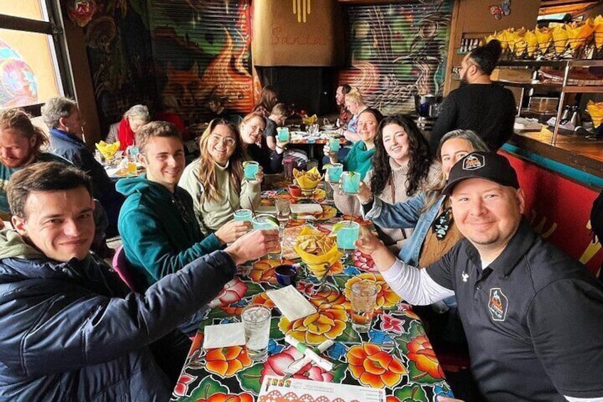 A group of people sitting around a table with colorful tablecloths and various items on it. 1
