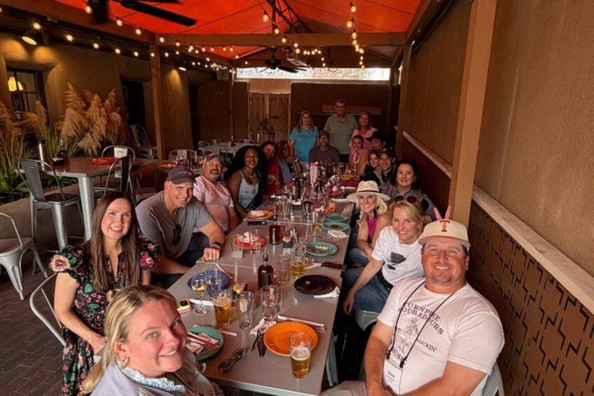 A group of people seated at an outdoor dining table with various dishes and drinks. 4