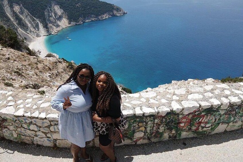 Deux personnes se tiennent près d’un mur de pierre, d’où elles dominent un paysage côtier avec des falaises et une plage. 3
