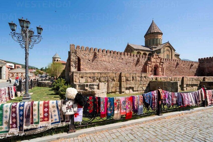 A historic stone wall with a church in the background, a street lamp, and a colorful cloth display. 3