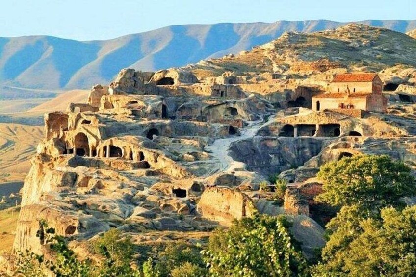 A rugged landscape with ancient rock-cut architecture, including caves and structures, set against a backdrop of mountains. 1