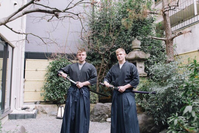 Deux personnes vêtues de costumes traditionnels, tenant des épées, se trouvent dans un jardin agrémenté de lanternes en pierre, avec un bâtiment en arrière-plan. 5