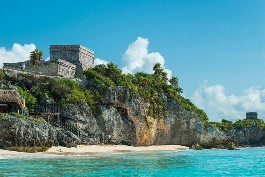 A coastal landscape with a large stone structure on a cliff, clear turquoise waters, and a sandy beach. 1