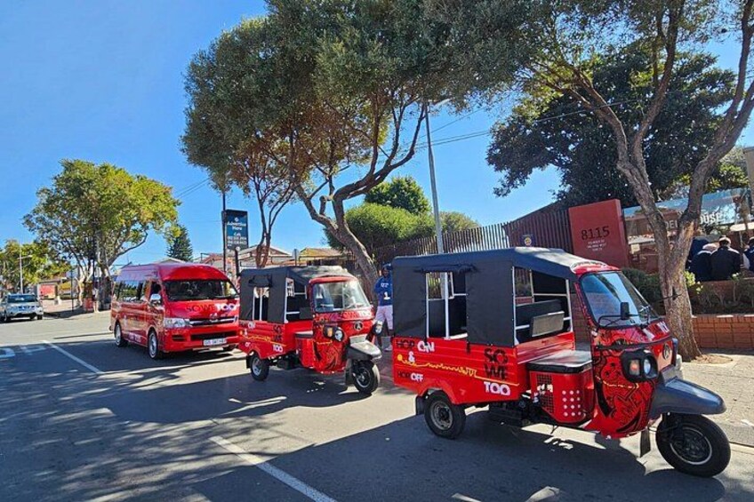 A street with tuk-tuks and a red van. Trees and a building with a sign are visible. 3