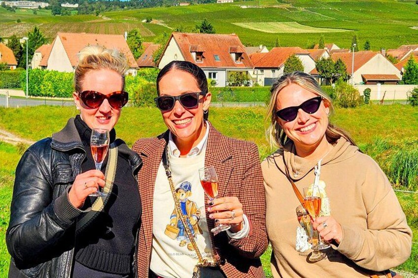 Tres personas sosteniendo copas de vino en un viñedo con casas al fondo. 2