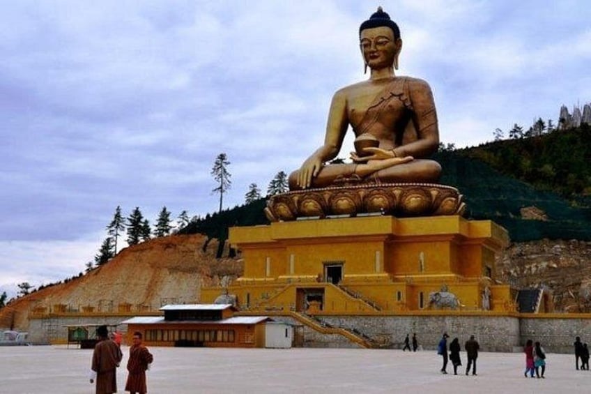 A large Buddha statue on a pedestal, surrounded by a few people and a building with a staircase leading up to it. 1