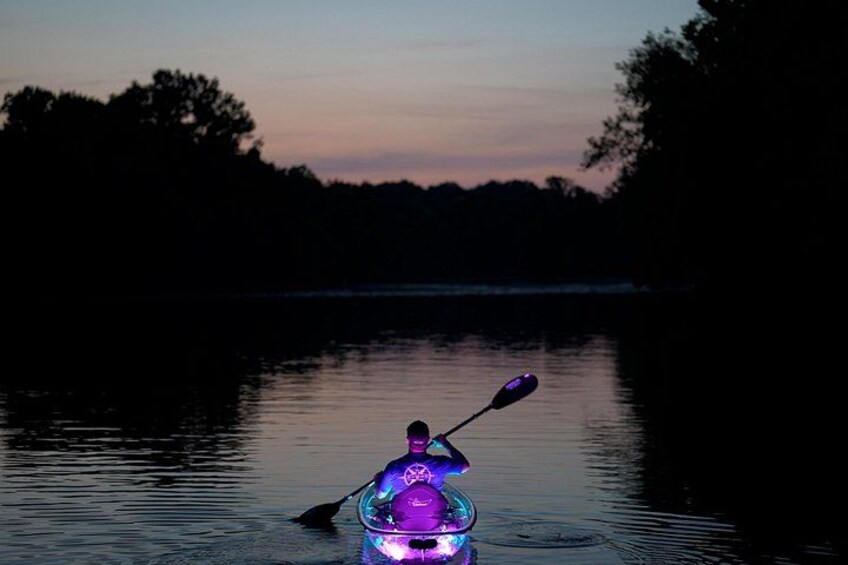 A kayak on calm water with a person paddling, surrounded by trees and a dusky sky. 1
