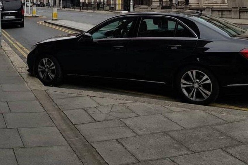 A black car parked on a city street with a sidewalk and buildings in the background. 3