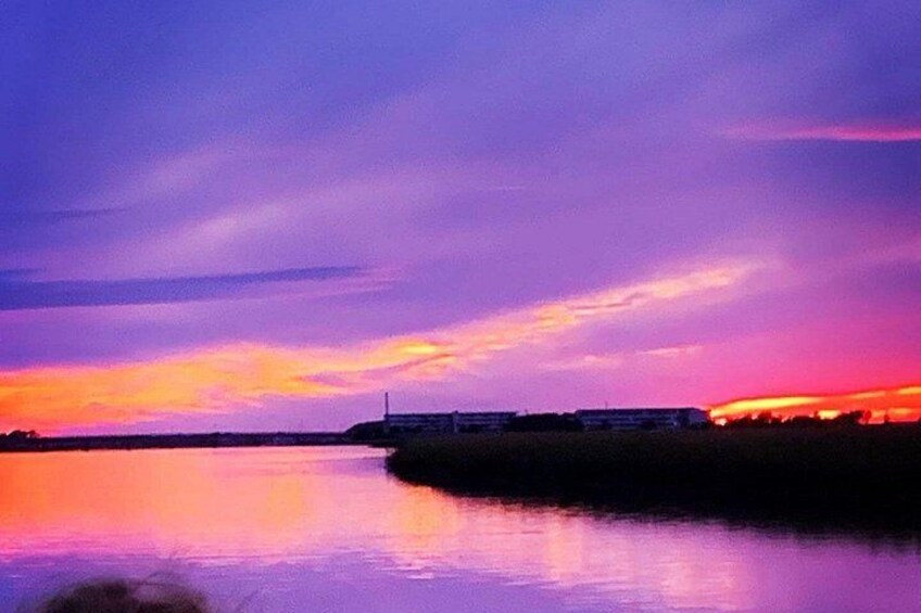 A sunset over a body of water with a distant shoreline and a building with lights on. 1