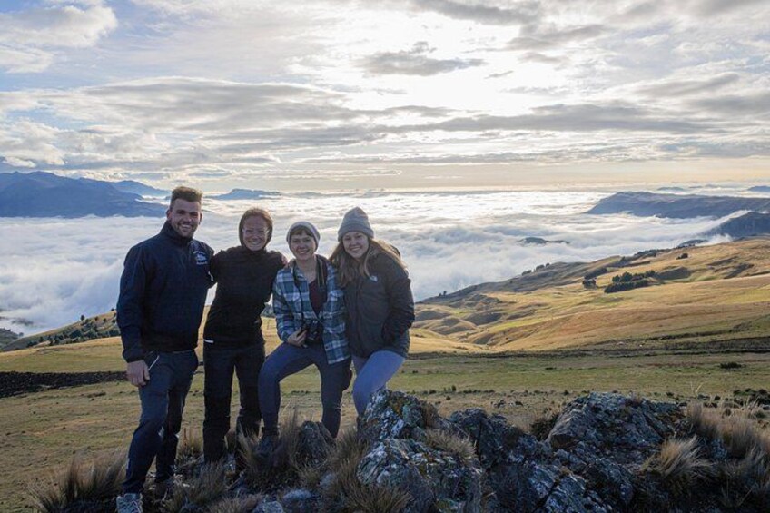 Cuatro personas de pie sobre una colina cubierta de hierba, con un paisaje montañoso y nubes al fondo. 4