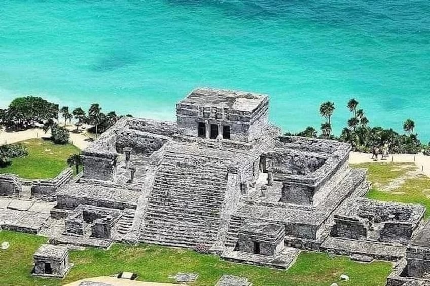 An ancient Mayan pyramid complex with a large central structure and smaller surrounding buildings, set against a turquoise sea and palm trees. 1